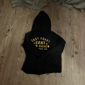 Svart hoodie från GANT - Säljer en svart hoodie från GANT med gul text 'EAST COAST NEW HAVEN' på framsidan. Tröjan har en klassisk design med huva och långa ärmar. Perfekt för en avslappnad stil.