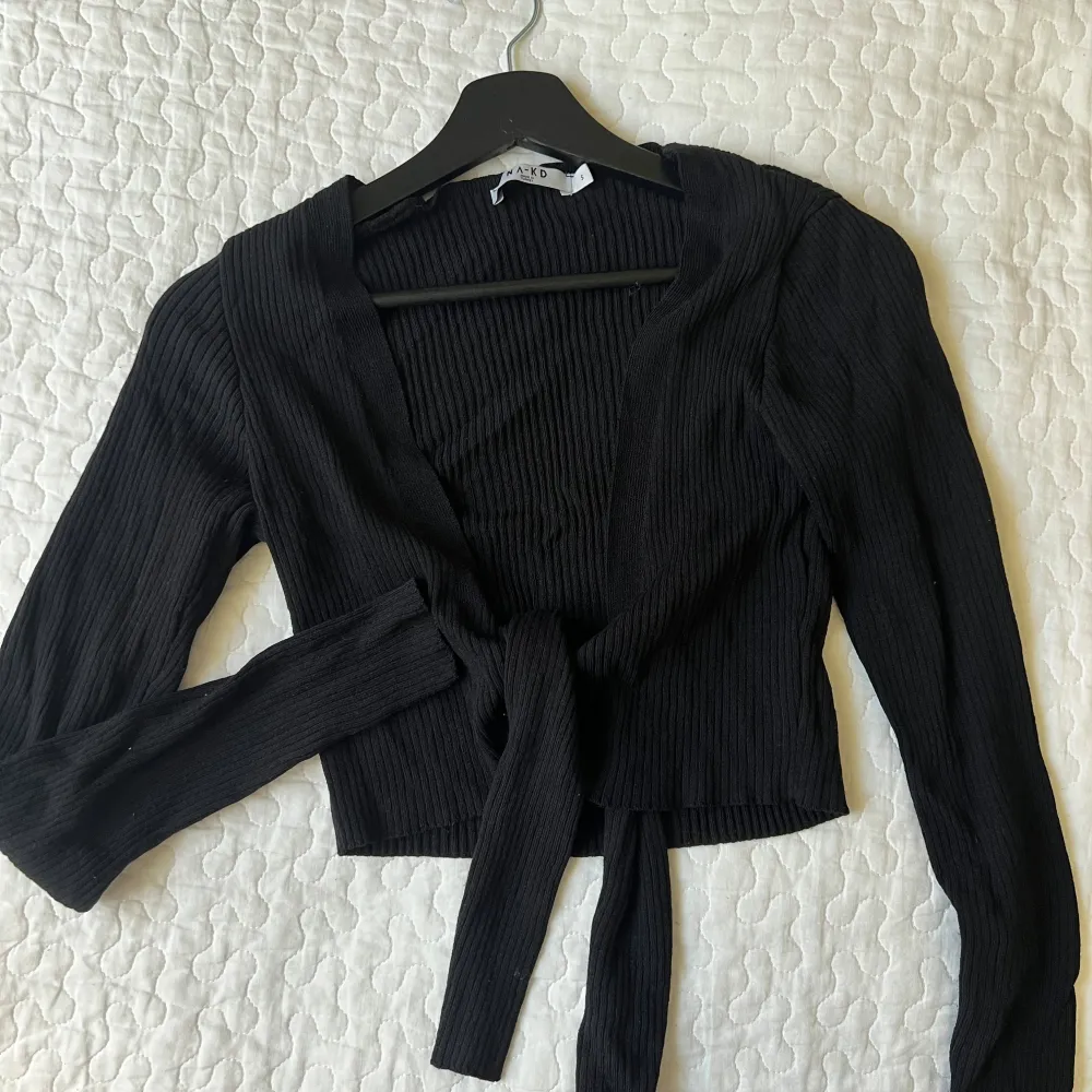 Snygg svart ribbad kofta med knytning framtill. Den har långa ärmar och en croppad passform, perfekt för lager på lager-stil. Ett stilrent plagg som funkar till många olika outfits.. Neuletakit & Villapaidat.