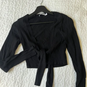 Svart ribbad kofta  - Snygg svart ribbad kofta med knytning framtill. Den har långa ärmar och en croppad passform, perfekt för lager på lager-stil. Ett stilrent plagg som funkar till många olika outfits.