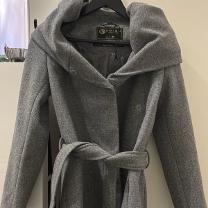 Grå kappa från Amisu - Snygg grå kappa från Amisu med stor huva och knytbälte i midjan. Perfekt för kyliga dagar med sin stilrena design och långa ärmar. Kappan har en elegant passform och är tillverkad i ett mjukt material.