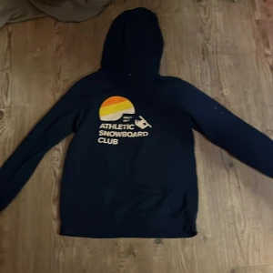 Mörkblå hoodie från Cubus - Säljer en mörkblå hoodie från Cubus med trycket 'Athletic Snowboard Club' i vitt och orange på framsidan. Tröjan har en klassisk huva och långa ärmar, perfekt för kyliga dagar. Materialet är en blandning av bomull och polyester för extra komfort.