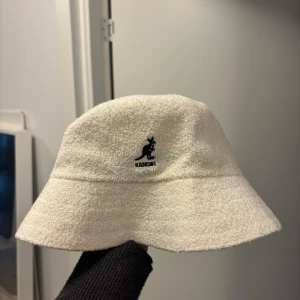 Vit bucket hat från Kangol - Snygg vit bucket hat från Kangol i modellen Bermuda. Perfekt för en avslappnad stil med sitt klassiska märke broderat framtill. Tillverkad i ett mjukt material som ger en skön passform.