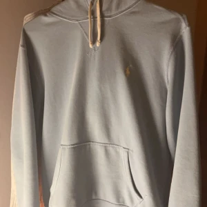 Ljusblå hoodie från Ralph Lauren - Säljer en stilren ljusblå hoodie från Ralph Lauren med klassisk känguruficka och justerbar huva. Den har en liten broderad logga på bröstet och är perfekt för en avslappnad look.