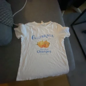 Vit t-shirt med tryck från H&M - Säljer en vit t-shirt från H&M med ett färgglatt tryck av apelsiner och texten 'California Oranges'. Perfekt för en avslappnad stil.
