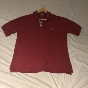  pikétröja från Burberry - Snygg röd pikétröja från Burberry med klassisk krage och korta ärmar. Tröjan har en diskret logga på bröstet och detaljer i det ikoniska Burberry-mönstret vid kragen. Perfekt för en stilren look.