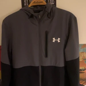 Grå och svart jacka från Under Armour - Snygg grå och svart jacka från Under Armour med dragkedja och huva. Perfekt för träning eller vardagsbruk. Jackan har en diskret logga på bröstet och en praktisk bröstficka med dragkedja.