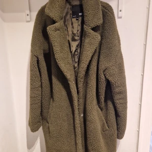 Olivgrön teddyjacka från 157 - Mysig olivgrön teddyjacka från 157 i storlek M. Jackan har en lång och avslappnad passform med långa ärmar och en mjuk, fluffig yta. Perfekt för kyliga dagar när du vill hålla dig varm och stilren.