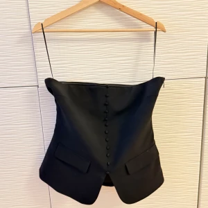 Svart tubtopp från Gina Tricot - Snygg svart tubtopp från Gina Tricot med knappar framtill och en liten slits. Fejkfickor som tillför en kostym-look. Perfekt för en stilren look. Toppen har en dragkedja på sidan för enkel på- och avtagning. Även en ”siliconlinje” runt bysten för att bättre fäste. Se bild med storleken. Storlek 40. Aldrig använd! Ny utan prislapp. Nypris 399 SEK. 