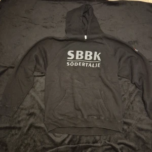 Svart hoodie med SBBK Södertälje-tryck - Snygg svart hoodie med stort SBBK Södertälje-tryck på bröstet. Tröjan har en klassisk känguruficka och en bekväm huva med dragsko. Perfekt för att visa ditt stöd för laget!
