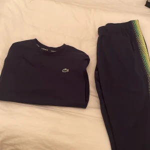 Mörkblå träningsset från Lacoste - Säljer ett snyggt mörkblått träningsset från Lacoste Sport. Tröjan har en klassisk passform med det ikoniska krokodilmärket på bröstet. Byxorna har elastisk midja och ett coolt mönster i grönt och gult längs sidorna. Perfekt för sportiga aktiviteter! storlek XS på Tröjan och storlek M på Byxorna.