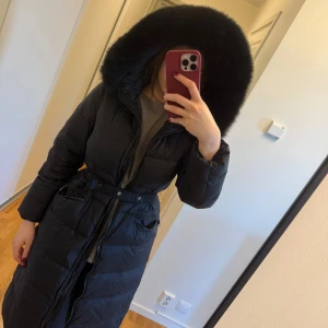 Svart lång dunjacka med pälsdetalj - Snygg svart lång dunjacka med pälsdetalj på huvan och bälte i midjan. Jackan har dragkedja framtill och långa ärmar, perfekt för kalla dagar. . 