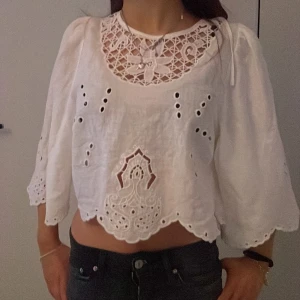 Vit blus med broderade detaljer - Charmig vit blus med broderade detaljer och vid passform. Blusen har trekvartsärmar och en vacker spetsdesign vid halsen. Den är croppad och har knytning i ryggen för en romantisk look. Från hm. Storlek S.
