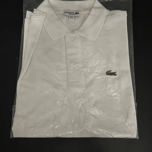 Vit pikétröja från Lacoste - Säljer en klassisk vit pikétröja från Lacoste med den ikoniska krokodilloggan på bröstet. Tröjan har en krage och knappar framtill. Perfekt för en stilren look.