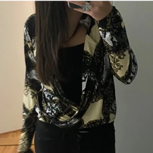 Mönstrad blus - Trendig blus med ett unikt mönster i svart, beige och guld. Blusen har långa ärmar och en omlott-design som ger en stilfull look. Perfekt för att ge din outfit en extra touch.💗