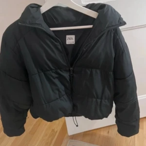 Svart pufferjacka från Zara - Snygg svart pufferjacka från Zara med dragkedja och hög krage. Jackan är croppad och har en oversized passform, perfekt för kyliga dagar. Den har långa ärmar och justerbar nederkant för extra komfort.