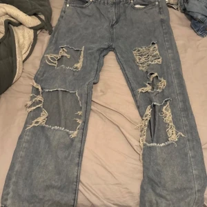 Blå jeans med slitningar - Säljer ett par blå jeans med coola slitningar på framsidan. De har en klassisk femficksdesign och stängs med dragkedja och knapp. Perfekta för en avslappnad stil.