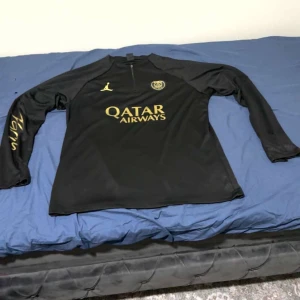 Svart fotbollströja och byxor från Jordan - Snygg svart långärmad fotbollströja och matchande byxor från Jordan. Tröjan har guldtryck med Qatar Airways och byxorna har Paris-tryck. Perfekt för träning eller casual wear.