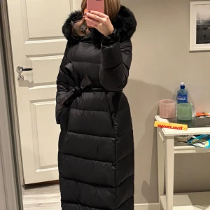 Svart lång dunjacka med pälskrage - Snygg svart lång dunjacka med en fluffig pälskrage och knytband i midjan. Jackan har långa ärmar och är perfekt för kalla vinterdagar men även kyligare vår och höstdagar. Den quiltade designen ger extra värme och stil. Jackan är köpt från Fur stockholm för 2700kr och är använd 1 och en halv vinter. Finns pyttelite smink vid kragen som knappt syns men går lätt att tvätta bort! 4e bilden är lånad!!