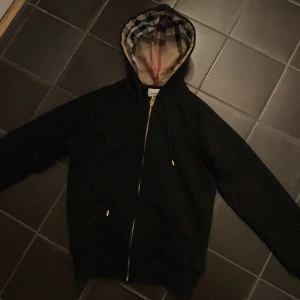 Svart hoodie med rutigt foder - Snygg svart hoodie från Burberry med dragkedja och klassiskt rutigt foder i huvan. Perfekt för en stilren look med en touch av lyx. Huvan har dragsko för justerbar passform.