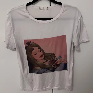Vit t-shirt med tryck - Säljer en vit t-shirt med ett färgglatt tryck av en tecknad figur som håller en Nutella-burk. T-shirten har korta ärmar och en rund halsringning. Perfekt för en avslappnad stil!