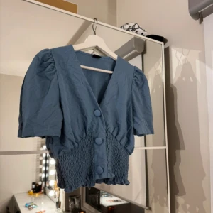 Blå blus med knappar - Charmig blå blus med korta ärmar och stora knappar framtill. Blusen har en smockad nederdel som ger en fin passform och volym vid axlarna för en trendig look.