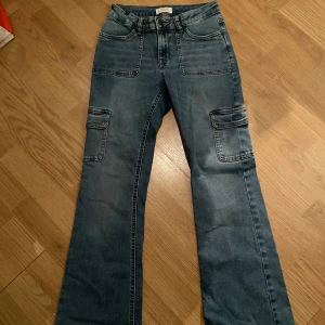 Blå cargobyxor från Lindex. - Snygga blå cargobyxor från Lindex med praktiska fickor på sidorna. Byxorna har en bootcut-stil och är perfekta för en avslappnad look. Tillverkade i jeansmaterial för en klassisk känsla.