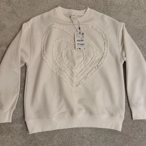 Vit sweatshirt med hjärtmönster - Vit sweatshirt från Zara med ett stort hjärtmönster i mitten. Tröjan har långa ärmar och en bekväm passform, perfekt för en avslappnad stil.