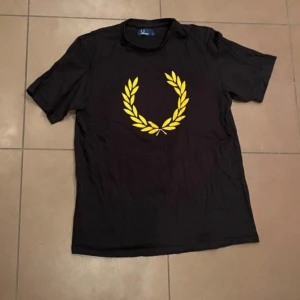 Svart t-shirt från Fred Perry - Säljer en svart t-shirt från Fred Perry med ett gult lagerkransmotiv på bröstet. Tillverkad i 100% bomull för en bekväm känsla. Perfekt för en stilren och avslappnad look.