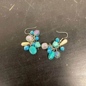 Färgglada örhängen och armband - Säljer ett set med färgglada örhängen och ett matchande armband. Örhängena har blå, turkosa och pärlemorskimrande pärlor i olika former. Armbandet har en tunn kedja med små färgglada detaljer. Perfekt för att liva upp vilken outfit som helst!