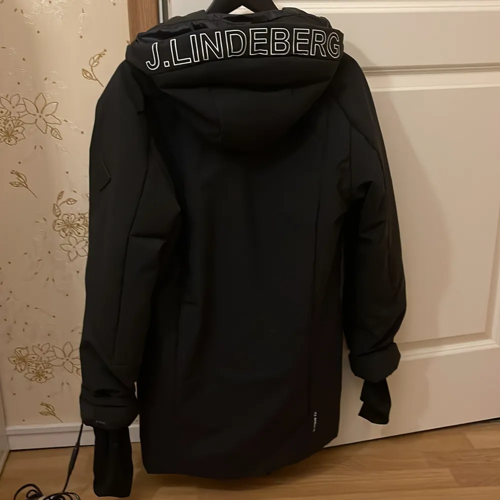 Snygg svart dunjacka från J.Lindeberg med dragkedja och huva. Jackan har en stilren design med logotypen tryckt på baksidan av huvan. Perfekt för kyliga dagar.. Takit.
