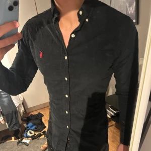 Ralph Lauren skjorta - Snygg svart skjorta i manchester från Ralph Lauren med klassisk krage och knappar framtill. Den har en röd broderad logotyp på bröstet och är långärmad. Perfekt för en stilren look.