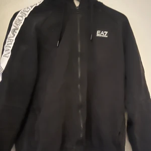 Svart hoodie från EA7 Emporio Armani - Snygg svart hoodie från EA7 Emporio Armani med dragkedja och huva. Den har en vit rand med logotyp längs ärmen och en liten logotyp på bröstet. Perfekt för en casual look.