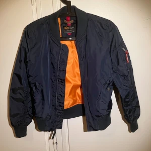 Blå bomberjacka från Alpha Industries - Snygg blå bomberjacka från Alpha Industries med orange foder. Jackan har en klassisk bomberstil med dragkedja och ribbade muddar. Perfekt för en cool och avslappnad look. Jackan har aldrig använts, jackan är i storlek 12a/152! (Nypris 1300kr)