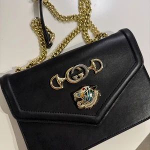 Svart axelväska ” INTE DEN RIKTIGA ” - Snygg svart axelväska med guldfärgad kedja och detaljer. Väskan har ett elegant GG-emblem och ett tigerhuvud på framsidan. Insidan har en dragkedja för extra säkerhet. Perfekt för att ge din outfit en lyxig touch.