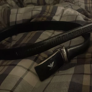 Svart Armani bälte - Snyggt svart bälte från Emporio Armani med en stilren silverfärgad spänne. Perfekt för att ge en elegant touch till din outfit. (Lite skråmor på skärmen av bältet)