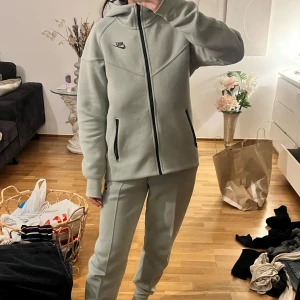Nike Tech Fleece mjukisdress - Säljer ett mjukisdress från Nike med dragkedja och huva. Tröjan har långa ärmar och praktiska fickor med dragkedja. Perfekt för en avslappnad look eller träning. Byxorna är moderna och sköna. Färgen är mer grön än vad som framkommer på bilderna, alltså mer lime grön än grå.