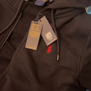 Svart hoodie från Ralph Lauren - Säljer en stilren svart zip hoodie från Ralph Lauren med dragkedja och röd broderad logga på bröstet. Perfekt för en avslappnad look. Hoodien har långa ärmar och en justerbar huva. Helt ny har bara använt 1 gång, etiketten följer med. Storleken är M men passar även S. Skick 10/10 (oanvänd) ✅ QR kod Scan funkar ✅ Pris kan diskuteras vid snabbaffär ✅