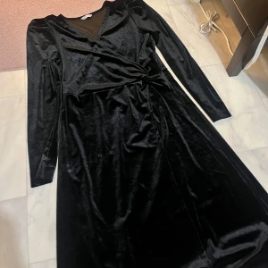 Svart sammet klänning - Elegant svart klänning i mjuk sammet med omlott-design och v-ringning. Den har långa ärmar och en knytning i midjan som ger en smickrande passform. Perfekt för en stilfull look.