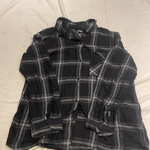 Svart och vit rutig skjorta - Säljer en svart och vit rutig skjorta med långa ärmar. Skjortan har en klassisk krage och knappar framtill. Perfekt för en avslappnad stil.