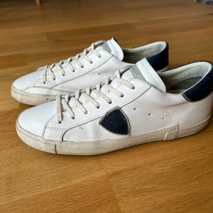 Philippe Model skor - Snygga vita sneakers från Philippe Model med mörkblå detaljer på häl och sula. Skorna har en klassisk design med snörning och en diskret logga på sidan. Perfekta för en stilren look. Har allt og plus kvitto köpta på farfetch. Skorna kommer slitna när man köper dem så dem är i bra skick. Fler bilder finns det är bara att fråga.