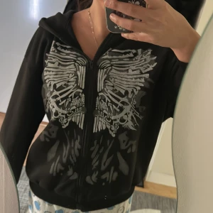 Svart hoodie med tryck - Säljer en svart hoodie med ett coolt tryck i silver på framsidan. Hoodien har dragkedja och långa ärmar, perfekt för en avslappnad stil. Den är både bekväm och stilren.