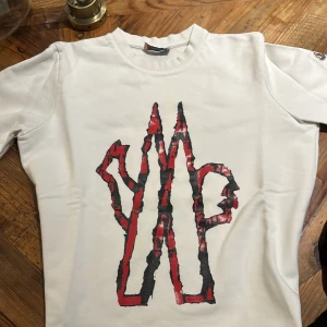 Vit t-shirt från Moncler med tryck - Snygg vit t-shirt från Moncler med ett coolt rött och svart tryck på framsidan. T-shirten har korta ärmar och Moncler-logga på ärmen. Perfekt för en avslappnad stil. Kan gå ner i pris med snabba affär