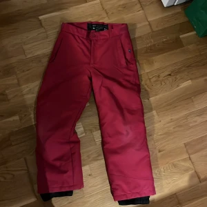 Röda skidbyxor med dragkedja - Snygga röda skidbyxor perfekta för vintern. De har en praktisk dragkedja och knappar framtill. Byxorna är designade för att hålla dig varm och bekväm i backen.