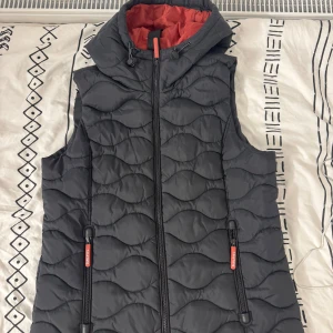 Svart quiltad väst från Sublevel - Säljer en svart quiltad väst från Sublevel med röd insida. Västen har en dragkedja framtill och en huva för extra skydd. Perfekt för kyligare dagar.