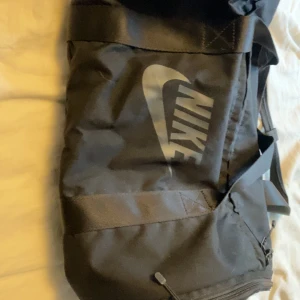 Svart sportväska från Nike - Säljer en svart sportväska från Nike med deras ikoniska logga på sidan. Väskan har en praktisk dragkedja och justerbar axelrem. Perfekt för träning eller som en rymlig weekendväska.aldrig använd ligger bara