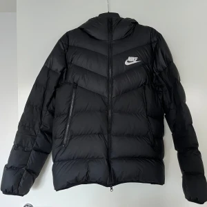 Dunjacka, Nike, M, svart - Svart dunjacka från NIKE i mycket fint skick, storlek M. Ifylld manuellt med äkta dun och fjädrar. Huva och dragkedja framtill samt fickor med dragkedja.   Mått: Längd ca 70 cm Bredd ca 55 cm Ärmlängd mätt från axeln ca 70 cm