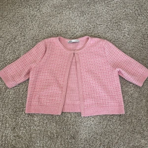 Rosa kofta från Zara - Säljer en söt rosa kofta från Zara med ett diskret rutmönster. Koftan har trekvartsärmar och en öppen design framtill. Perfekt för en stilren och bekväm look.