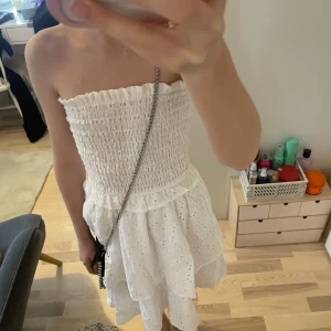 Vit axelbandslös klänning - Säljer en söt vit axelbandslös klänning med volanger och broderade detaljer. Perfekt för sommarens alla tillfällen. Klänningen har en smockad överdel som ger en bekväm passform.