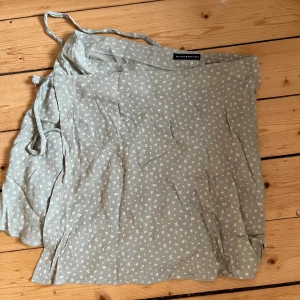 Grön omlottkjol från Brandy Melville - Söt grön omlottkjol från Brandy Melville med små vita blommor. Kjolen har knytband i midjan och en lätt volangkant. Perfekt för en avslappnad och stilren look. 