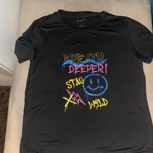Svart t-shirt från SHEIN med färgglatt tryck - Cool svart t-shirt från SHEIN med färgglatt tryck i blått, rosa och gult. Texten 'Breathe Deeper! Stay Wild' och en glad smiley ger en lekfull touch. Perfekt för en avslappnad stil.